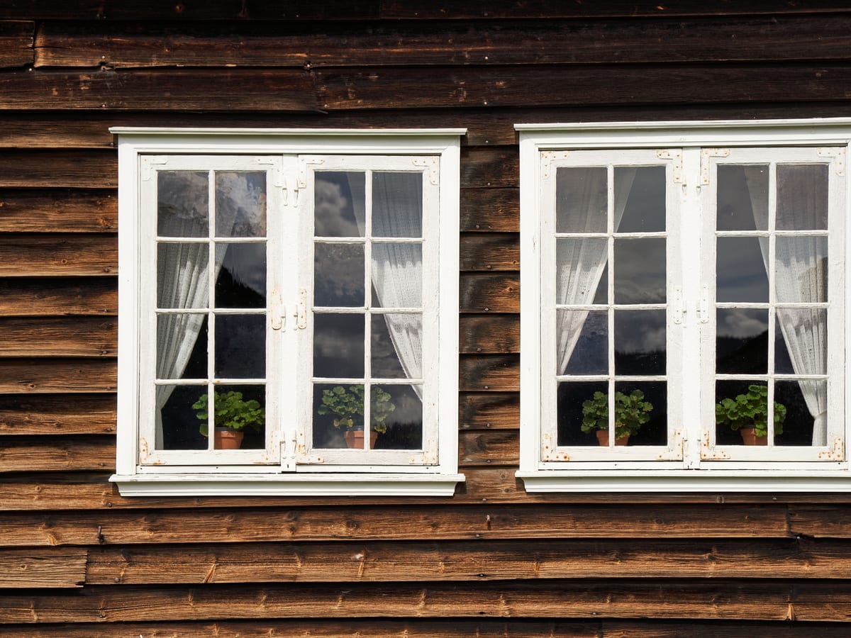 Close-up of two white framed windows with curtains against a rustic wooden house exterior.